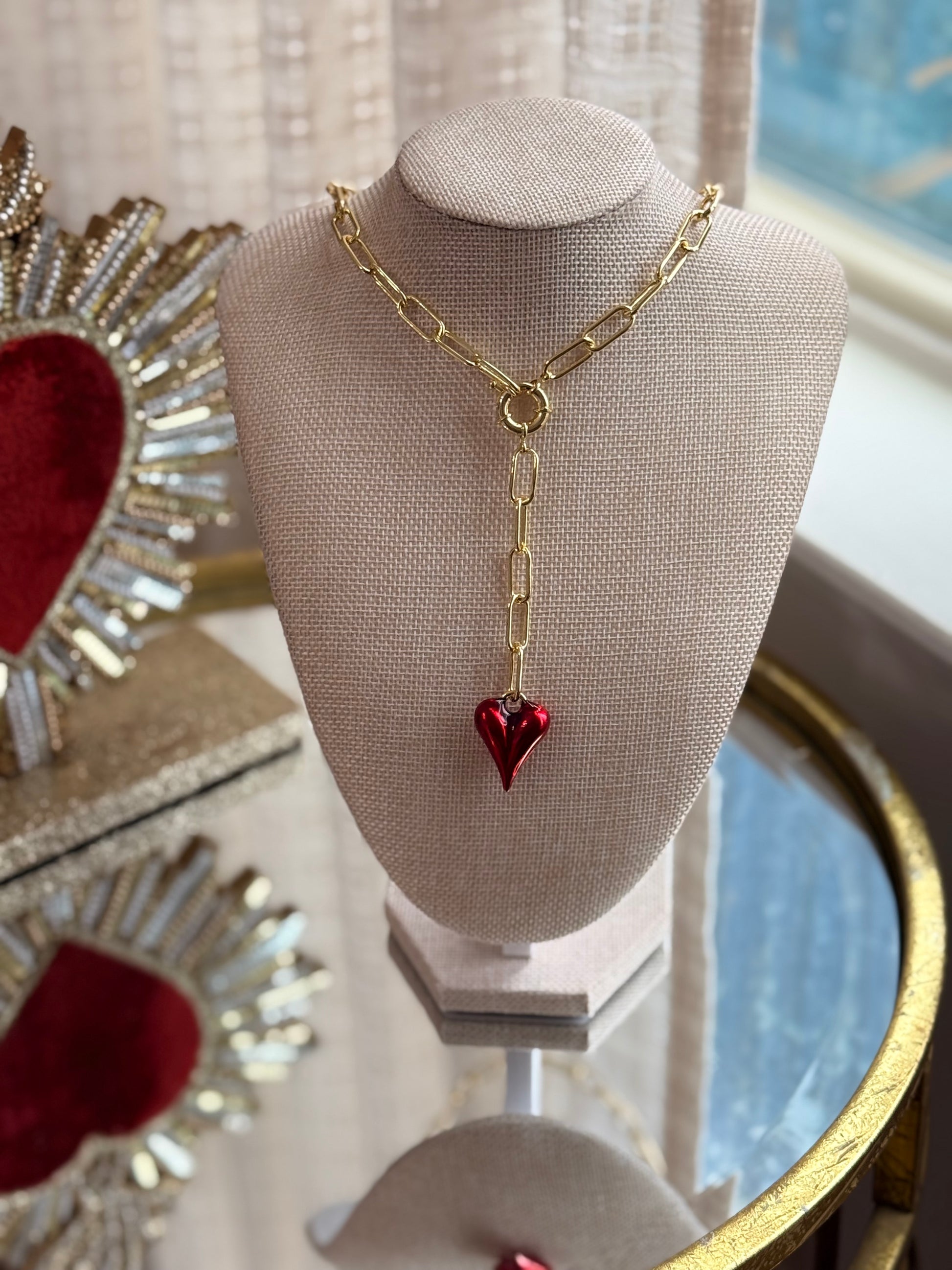 Gold necklace with a red heart pendant on a mannequin bust, with decorative items in the background.