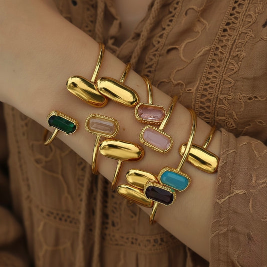 Gold bracelets with colorful gemstones on a wrist against a brown lace fabric background
