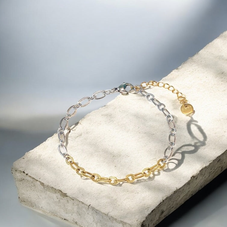 Two chain bracelets, one gold and one silver, on a stone surface with a blurred background.