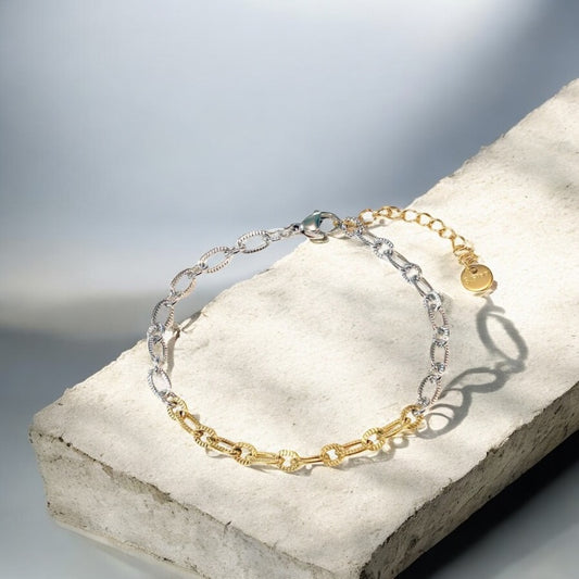 Two chain bracelets, one gold and one silver, on a stone surface with a blurred background.